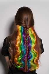 Close-up shot of a brown-haired girl with multicolored strands of hair. Colored clip in hair extensions. The woman in the black top with rainbow tresses is on a gray background. Back view.