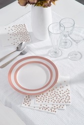 Close-up of white and pink plates, three glasses, white napkins with rose gold dots, eating utensils and a vase of flowers on the table with a white tablecloth. Top view.
