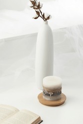 Still photo of an aromatic colored candle adorned with a cord and a key. The designer handmade candle is located on a board against a wall. There is a vase with flowers and a book on the table.       
