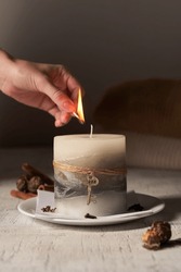 Cropped close-up shot of a woman's hand lighting an aromatic colored candle adorned with a cord and a key on a grey background. The designer handmade candle is located on a napkin on a white plate.   