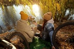 a guy and a girl on a bike ride sat on a blanket near the water and drink tea from a thermos