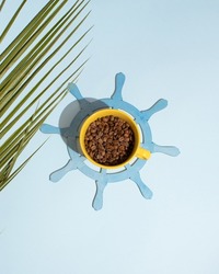Yellow mug filled with coffee beans stands on the rudder on bright blue background decorated with palm leaf shadow. Minimal summer brake concept. Creative tropical refreshing beverage idea.