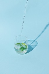 Pouring drink in clear glass with mint leaves inside on bright blue background. Minimal refreshing summer beverage concept. Lemon soda or tropical cocktail drink idea.