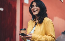 Happy girl in hipster eyeglasses listening to music while going to work, cheerful hispanic woman drinking coffee outdoors using modern smartphone device, happy smiling european businesswoman walking