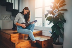 Beautiful young woman wearing casual clothes enjoying play ukulele guitar in the morning sitting on wooden stairs at home near big window, Flare Light, Happy lifestyle Leisure Activities Concept