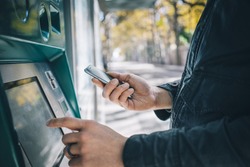 Closeup of male hands using smart phone while typing on ATM, bank machine