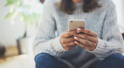 Close-up image of young hipster girl sitting at cozy home interior and using modern smartphone device, female hands typing text message via cellphone, social networking concept