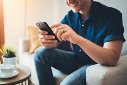 Happy smiling young man using modern smartphone device while sitting on sofa at home, modern design interior, cheerful hipster guy typing an sms message at social network