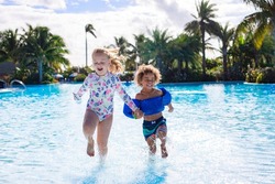 A Cute diverse boy and little girl running and splashing together holding hands in the large swimming pool resort while on a family vacation