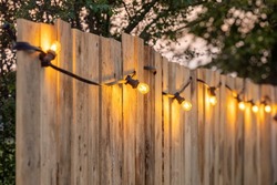 Cosy light bulbs lined up in a row, against a wooden garden fence. There are some green bushes and green grass in the background. It's a sunny Summer day evening at a country wedding or other