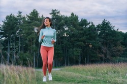 An astonishing young woman is jogging with a thick green forest behind her, listening to music on her smart phone and enjoying the weather.