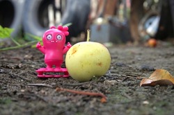 A small toy monster, pink plastic alien on the ground near green apple. Close-up photo on the background of tires in the garden.