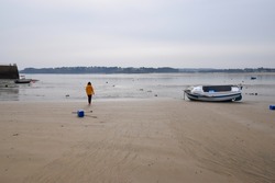 Woman in bright yellow jacket walking on a sandy beach towards the sea at low tide, with small boats moored in the harbor
