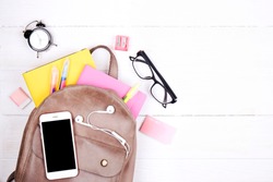 Hipster grey leather backpack, full of school supplies, blank screen cell phone, earphones, pink & yellow notebook, glasses, mini alarm clock. Back to school concept. Close up, copy space, flat lay.