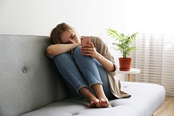 Portrait of beautiful young woman with depressed facial expression sitting on grey textile couch holding her phone. Cyber bullying victim concept. Sad female in her room. Background, copy space.