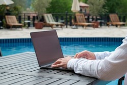 Young man wearing white linen shirt working on his laptop by the pool. Male travel blogger typing on keyboard. Perks of being a freelancer. Close up, copy space, background.