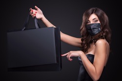 Young woman in a medical mask showing on bag in black Friday holiday. Sale and shopping concept in 2020 - time of coronavirus pandemic.  