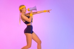 Emotional fit woman in sportswear screaming using megaphone. Social issues, protest concept. Side view. A girl speaks into a megaphone about an upcoming promotion and discounts in fitness store