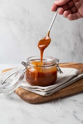 Homemade salted caramel sauce in glass jar on wooden board, grey background. Caramel sauce drips from spoon in hand.