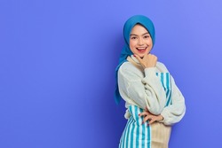 Portrait of smiling young housewife woman in hijab and apron looking at camera, pointing aside with finger isolated on purple background. People housewife muslim lifestyle concept