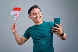 Excited young Asian man holding mobile phone and indonesian flag while celebrating indonesia independence day isolated on grey background. indonesia independence day celebration concept