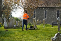 Man wearing a high visibility orange jacket tends the local churchyard keeping the grass cut around ancient gravestones with a motor mower. Arched windows of the church in the background.