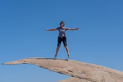 Potato Chip Rock, Ramona, California; a natural granite formation with wafer-thin outcrop leads into a clear blue sky with copy space. An older caucasian woman star jumps on a fragile looking overhang