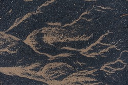 Random natural patterns made by water retreating across dark gritty sand in a river estuary. The texture is black and tan with the detail running horizontally. Small stones and grit dot the surface.