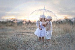 Happy and inspired angels boy and girl stand in the pouring rain hiding under an umbrella. A rainbow is visible in the sky. Thin streams of rain are barely visible on the whole background. 
