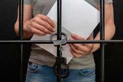 A handcuffed prisoner behind bars holds a white sheet of paper in his hand.  Concept: petition for pardon, court verdict, defense without a lawyer.