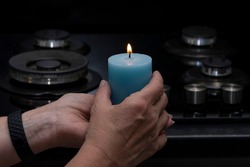 An elderly woman holds a burning candle in her hands, against the background of a non-working gas stove, selective focus. Concept: turning off heating and gas, reducing room temperature.