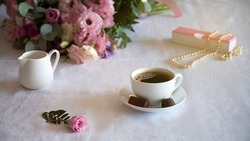 Bouquet of gentle pink flowers on the table with white tablecloth . White cup with coffee and chocoltae.Pearl necklace on the gift box. Top view. concept of breakfast in cafe.