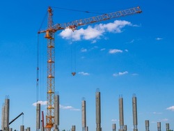 A tower construction crane on the background of a blue sky with clouds. Boom rotary crane with boom. Reinforced concrete piles and frame during the construction of a multi-storey building.