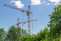 Tower construction cranes and trees against a blue sky with white clouds. Boom rotary crane with boom. Construction of apartment buildings in the countryside. Construction of high-rise buildings.