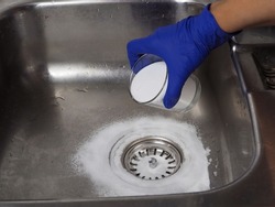 Gloved hand putting baking soda on drain in kitchen sink from glass jar. Close up. Eco friendly house cleaning concept.