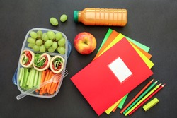 School supplies and lunch box with tasty rolls, cucumbers, carrots, grapes, apple, bottle of juice on black background, back to school concept. Horizontal. Top view.