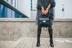 close up accessories details of stylish woman walking in city in warm fur coat, winter season, cold weather, holding leather handbag, legs in boots, footwear street fashion trend