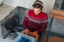 young attractive serious concentrated man sitting on sofa at home in winter, wearing red knitted sweater, working on laptop, freelancer, busy, listening to headphones, typing, studying online