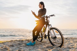 attractive slim woman riding bicycle, sport on morning sunrise beach in fitness sportswear, healthy lifestyle, listening to music on wireless earphones holding smartphone, relaxing smiling happy