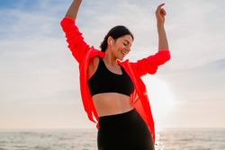 young smiling attractive slim woman doing sports in morning sunrise dancing on sea beach in sports wear, healthy lifestyle, listening to music on earphones, wearing pink windbreaker jacket, having fun