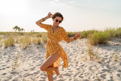 stylish attractive slim smiling woman on beach in summer style fashion trend outfit carefree and happy, feeling freedom, wearing yellow printed dress boho style chic and sunglasses