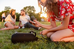 young hipster company of friends having fun together in park smiling listening to music on wireless speaker connecting to smartphone, summer style season