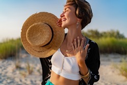 stylish attractive slim smiling woman on beach in summer style fashion trend outfit happy having fun wearing white top and tunic boho style chic and straw hat