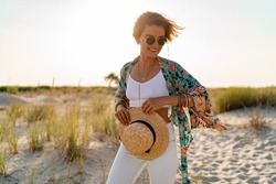 pretty attractive slim smiling woman on sunny beach in summer style fashion trend outfit happy, freedom, wearing white top, jeans and colorful printed tunic boho style chic and straw hat