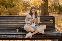 candid attractive young smiling woman sitting on bench in autumn park using phone wearing checkered coat, happy mood, fashion style trend