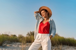 stylish attractive slim smiling woman on beach in summer style fashion trend outfit happy having fun wearing white pants red top blue shirt boho style chic and straw hat