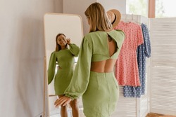 young pretty woman in green dress trying on fashion style trend dress looking in mirror at home or showroom