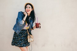 young beautiful happy stylish hipster girl, cocktail, smoozy drink, denim jacket, smiling, fashion, teen, cool accessories, purse, hat, sunglasses, amazed, vintage style, wall background, hair, wind