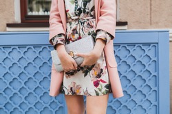 young beautiful stylish woman walking in street in pink coat, floral printed dress, holding silver purse in hands, autumn fashion trend, hands close-up, details