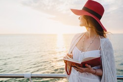 young beautiful woman walking by the sea, reading travel diary book, morning sunrise, romantic mood, red hat, thinking of memories, bohemian style outfit
